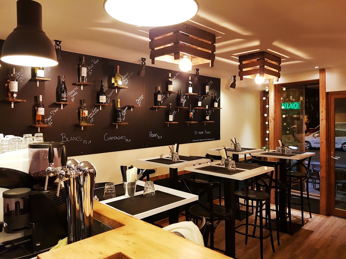 Salle du Cosy avec mur noir habillé d'étagères de bouteilles, tables hautes blanches et bois clair.